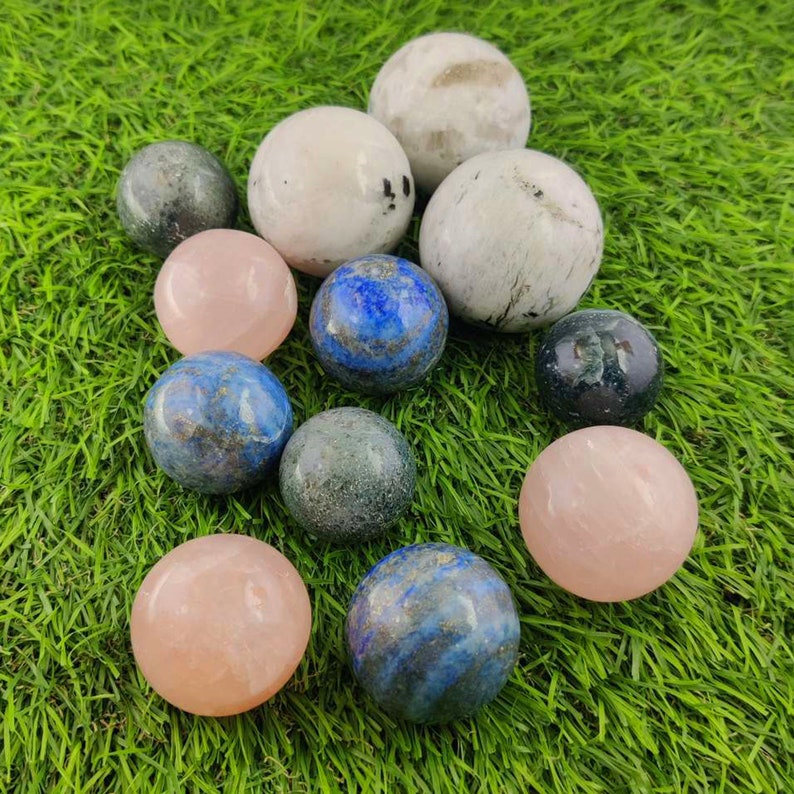 Pu&ograve; includere: Una collezione di sfere di pietre preziose lucidate in vari colori, tra cui rosa, blu, verde e bianco. Le sfere sono disposte su un letto di erba verde.
