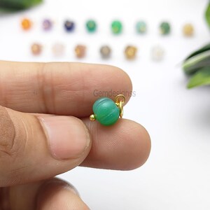 May include: A small, green, round gemstone charm with gold-colored accents. The charm has a ribbed texture and a small gold loop for attaching to jewelry. The background features other colorful gemstones.