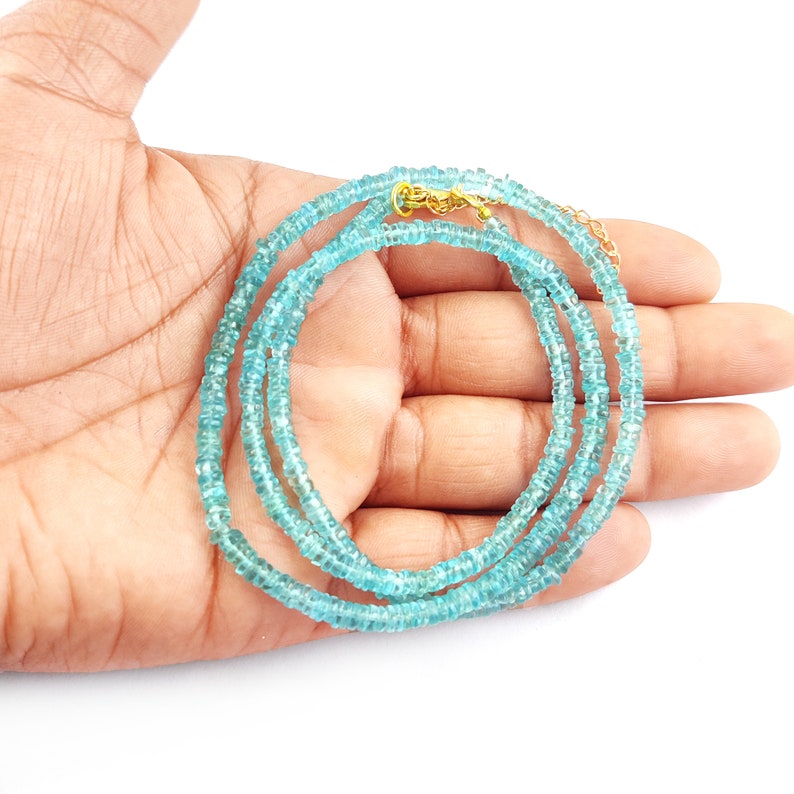 May include: A delicate necklace composed of small, light blue beads. The necklace is displayed against a white background, with a gold-colored clasp. The beads are evenly spaced, creating a refined and elegant appearance. The necklace is approximately 18 inches long.