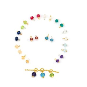 Könnte beinhalten: Ein Set mit 12 Edelstein-Anhängern mit goldenen Sprungringen. Die Anhänger sind in einem Regenbogen von Farben, darunter Rot, Orange, Gelb, Grün, Blau, Indigo und Violett. Die Anhänger sind in einem Kreis angeordnet, wobei die goldenen Sprungringe nach außen zeigen.