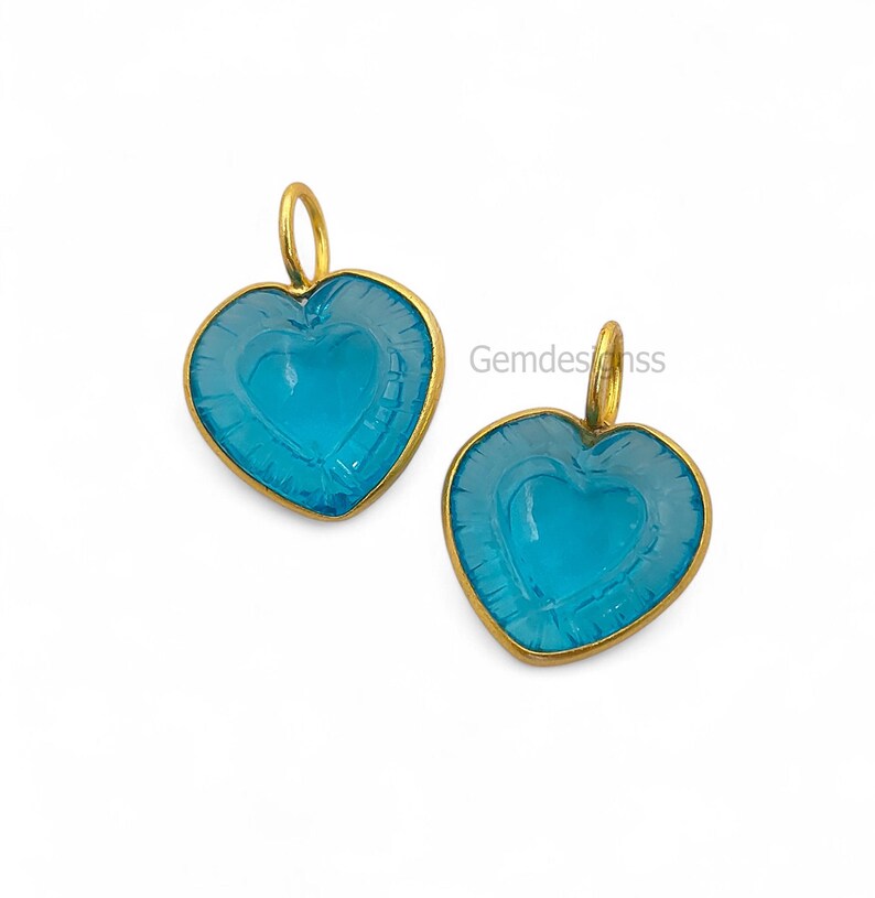 May include: Two heart-shaped pendants with a light blue centre and a gold-coloured frame. The pendants have a loop at the top for hanging. The text "Gemdesignss" is visible on the right side.