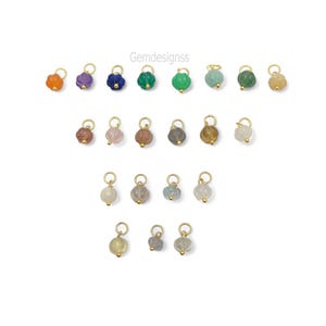 May include: A collection of gemstone charms in various colors, including orange, purple, blue, and green. Each charm has a gold-colored loop and a small gold bead. The charms are arranged on a white background. The text "Gemdesignss" is in the upper right corner.