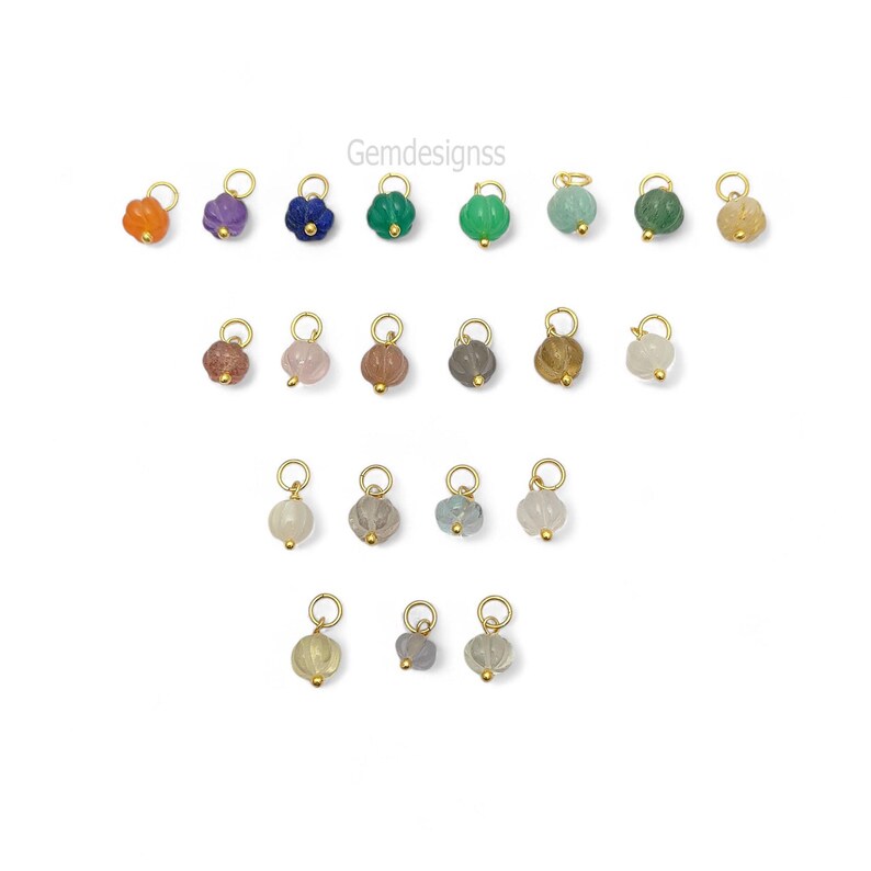 May include: A collection of gemstone charms in various colors, including orange, purple, blue, and green. Each charm has a gold-colored loop and a small gold bead. The charms are arranged on a white background. The text "Gemdesignss" is in the upper right corner.