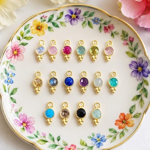 May include: Sixteen small gold-toned jewelry charms, each featuring a circular faceted gemstone in various colors including pink, blue, green, purple, and black, with three tiny gold spheres at the base and a small loop at the top for attachment.