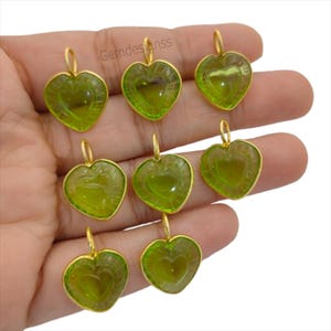 May include: Nine heart-shaped pendants with a gold-coloured frame and a green centre. Each pendant has a loop at the top for hanging. The pendants are arranged in a hand.