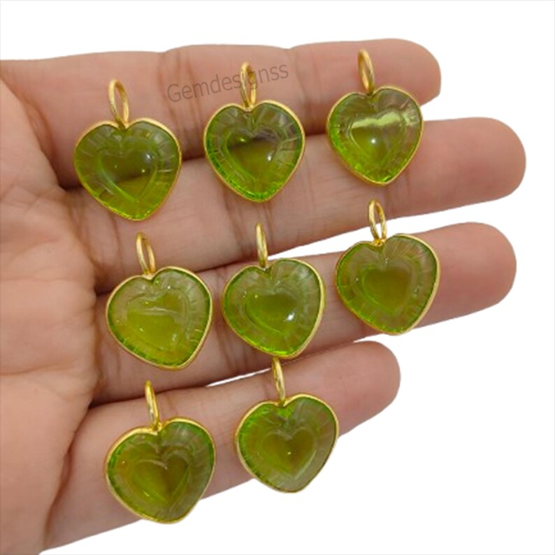 May include: Nine heart-shaped pendants with a gold-coloured frame and a green centre. Each pendant has a loop at the top for hanging. The pendants are arranged in a hand.