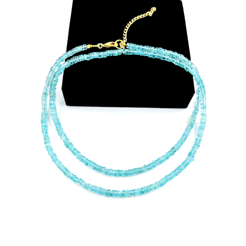 May include: A delicate, double-strand necklace featuring small, light blue beads. The necklace has a gold-colored clasp and chain. The jewelry is displayed against a black background and a white surface.