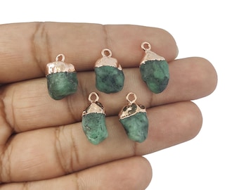 May include: Five raw emerald pendants with rose gold-colored caps and loops. The emeralds are a deep green color and have a natural, rough texture. The pendants are displayed against a neutral background.