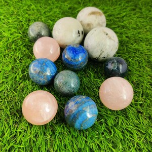 Pu&ograve; includere: Una collezione di 11 sfere di pietre preziose lucidate in vari colori, tra cui rosa, blu, verde e bianco. Le sfere sono disposte su una superficie verde erbosa.