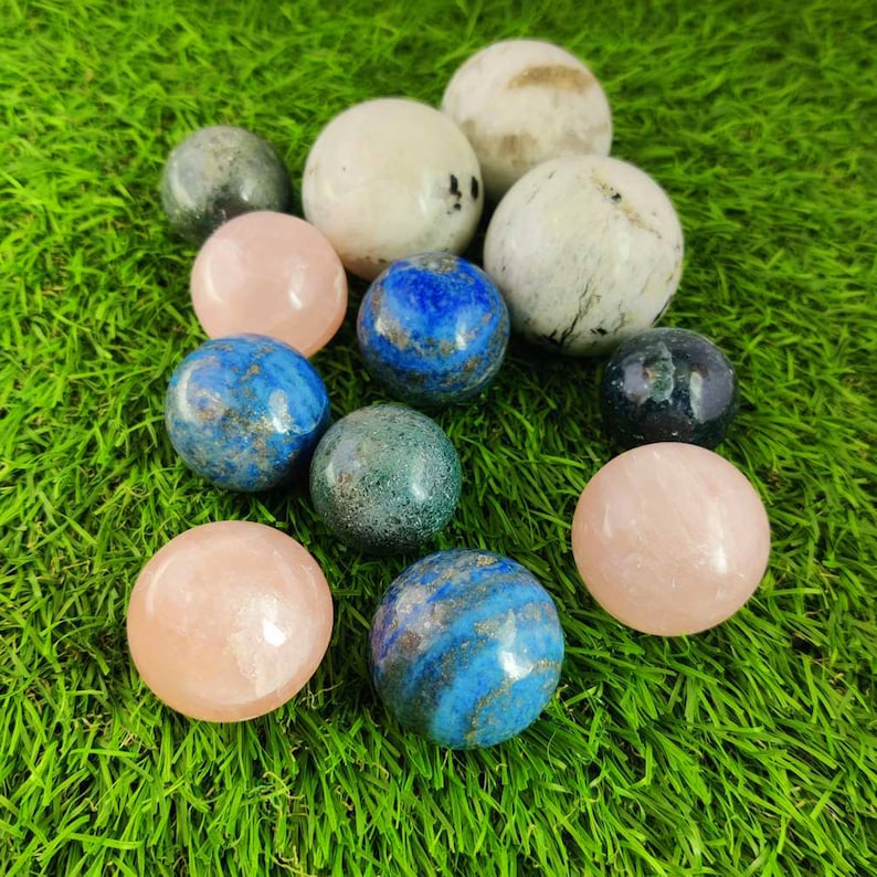 Pu&ograve; includere: Una collezione di 11 sfere di pietre preziose lucidate in vari colori, tra cui rosa, blu, verde e bianco. Le sfere sono disposte su una superficie verde erbosa.