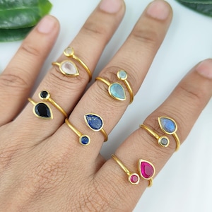 May include: A set of six gold rings with different colored gemstones. Each ring features a teardrop-shaped gemstone and a smaller round gemstone. The gemstones are set in gold and the rings are adjustable.