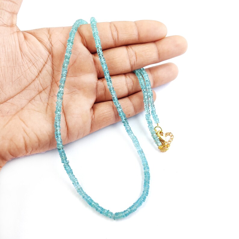 May include: A delicate necklace featuring small, light blue beads. The necklace has a gold-colored clasp and is displayed on a white background. The beads appear to be evenly sized and strung together to form a necklace.