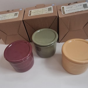 May include: Three ceramic storage jars with lids in different colors: a green jar with a green lid, a maroon jar with a maroon lid, and a yellow jar with a yellow lid. The jars are sitting on a white surface.