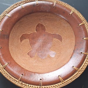 May include: A round, brown decorative bowl with a turtle carving in the center. The bowl has a woven, light brown rim and is stitched with gold-colored thread. The turtle carving is a lighter shade of brown than the bowl's rim.