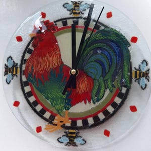 May include: A round glass clock with a rooster design. The rooster is red, green, and blue with a black and white checkered border. The clock has black hands and a gold center. There are bees around the edge of the clock.