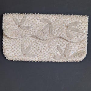 May include: A white beaded clutch purse with a scalloped edge and a floral design. The purse is covered in small white beads and sequins.