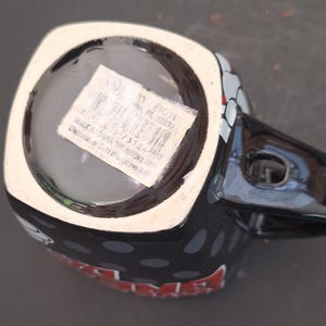 May include: Black ceramic mug with a handle, featuring a design with white, red, and gray accents. The mug has a white rim and a black interior. The bottom of the mug has a sticker with text and a barcode.