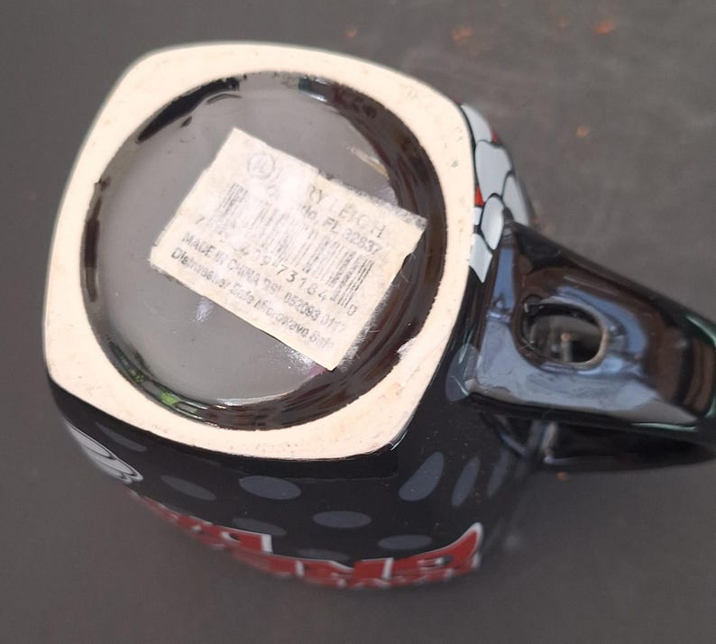 May include: Black ceramic mug with a handle, featuring a design with white, red, and gray accents. The mug has a white rim and a black interior. The bottom of the mug has a sticker with text and a barcode.