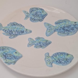 May include: A white ceramic plate decorated with six blue and green fish. The fish are arranged in a circular pattern, with varying sizes and orientations. The plate has a slightly raised edge.