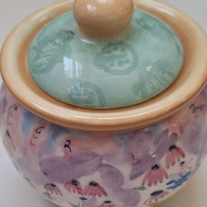 May include: A hand-painted ceramic jar with a lid. The jar features a floral design in shades of purple, pink, and blue. The lid is light green with a beige knob. The rim of the jar is beige.