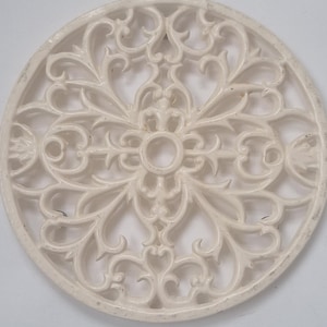 May include: Off-white, circular trivet with an ornate, openwork design. The trivet features a repeating pattern of swirling lines, floral motifs, and a central circular opening. The edges are slightly raised, and the overall aesthetic is vintage or antique.