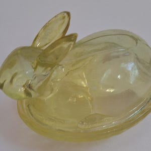 May include: A yellow glass rabbit-shaped butter dish with a lid. The dish is made of clear glass with a slight yellow tint. The rabbit has long ears and a rounded body.
