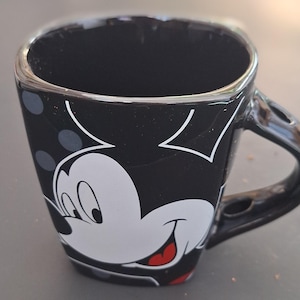 May include: Black square-shaped coffee mug featuring a white Mickey Mouse face with a red tongue and a black handle. The mug has a black background with white polka dots and white Mickey Mouse ears.