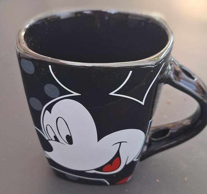 May include: Black square-shaped coffee mug featuring a white Mickey Mouse face with a red tongue and a black handle. The mug has a black background with white polka dots and white Mickey Mouse ears.