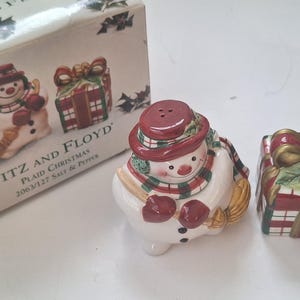 May include: A ceramic salt and pepper set featuring a snowman and a gift box. The snowman wears a red hat and scarf, holding a broom. The gift box is red and green with a gold bow. The box reads "FITZ AND FLOYD PLAID CHRISTMAS".