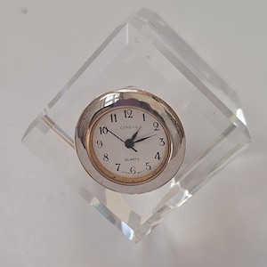 May include: A clear, diamond-shaped acrylic clock holder with a silver-colored clock face. The clock face is white with black numerals and hands, and the word "GENEVA" is printed above the center. The clock reads 2:10.