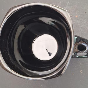 May include: A glossy black ceramic mug with a unique square-like shape and a handle. The interior is black with a white circle at the bottom. The mug has a shiny, reflective surface.