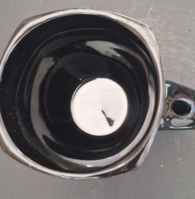 May include: A glossy black ceramic mug with a unique square-like shape and a handle. The interior is black with a white circle at the bottom. The mug has a shiny, reflective surface.