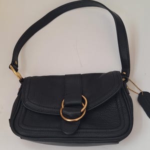 May include: Black leather shoulder bag with a flap closure and a gold-tone metal ring detail. The bag has a single shoulder strap and a small tag attached. The bag is made of textured leather.