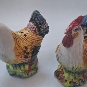 May include: A pair of ceramic rooster and hen salt and pepper shakers. The rooster has a red comb and a white body with brown and yellow feathers. The hen has a white body with brown and yellow feathers. Both are perched on a green base.