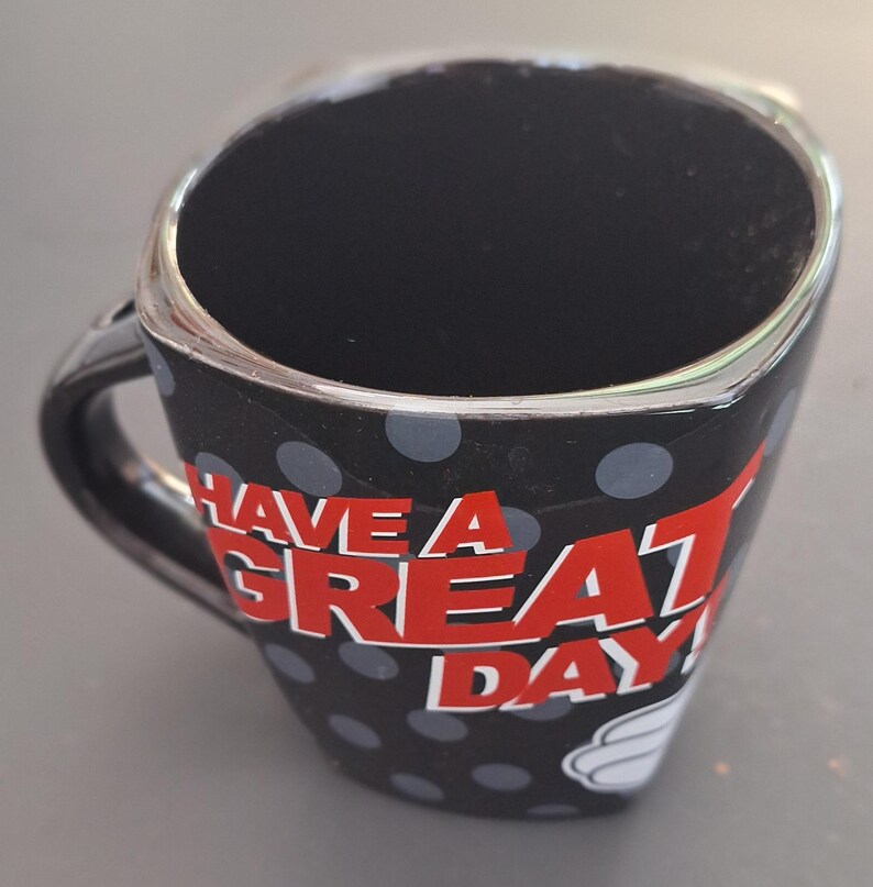 May include: Black ceramic mug with a silver rim and handle, featuring a polka dot pattern with gray dots. The mug has the text "HAVE A GREAT DAY!" in red and white. A white glove design is also visible.