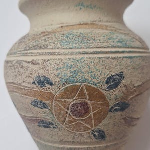 May include: A small, textured ceramic vase with a beige base color. The vase features a decorative design with a star and leaf pattern in shades of brown, blue, and red. The vase has a rounded body and a narrow neck.