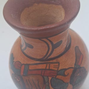May include: A small, round, terracotta-colored vase with a wide, flared rim. The rim and interior are a darker brown. The vase is decorated with black and red painted figures and designs. A unique piece of pottery.