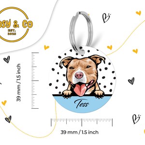 Puede incluir: Una chapa redonda blanca con lunares negros. Una imagen de dibujos animados de un perro pit bull marr&oacute;n y blanco con la lengua rosa que sobresale est&aacute; mirando por encima de la parte superior de la chapa. La chapa tiene un fondo azul con el nombre "Tess" escrito en blanco. La chapa est&aacute; unida a un llavero plateado.