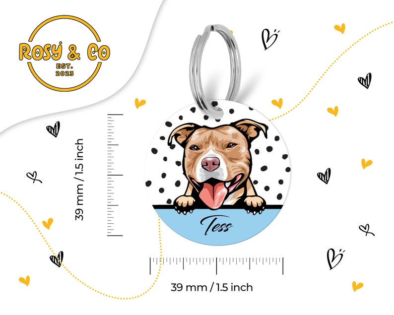 Puede incluir: Una chapa redonda blanca con lunares negros. Una imagen de dibujos animados de un perro pit bull marr&oacute;n y blanco con la lengua rosa que sobresale est&aacute; mirando por encima de la parte superior de la chapa. La chapa tiene un fondo azul con el nombre "Tess" escrito en blanco. La chapa est&aacute; unida a un llavero plateado.