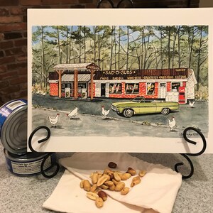 May include: Watercolor painting of a roadside store named "SAC-O-SUDS" with a gas pump, a green convertible, and chickens. The store sells "PNUTS BAR-BQ WINE BEER GROCERIES COLEDRINKS ENFTACK". The artwork is displayed on a black metal stand.