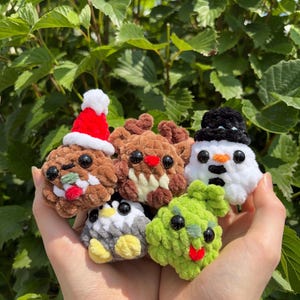 May include: A collection of five small, plush, crocheted figures featuring a brown creature with a red Santa hat, a brown reindeer-like figure, a white snowman with a black hat, a black and white penguin, and a bright green textured creature.