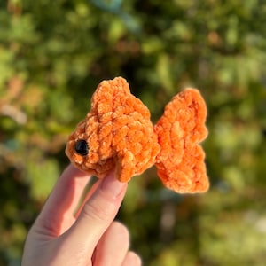 May include: A handmade, orange crocheted goldfish with a black bead eye. The plush fish has textured, fluffy details and is held up against a blurred green background. This soft toy is a unique, handcrafted item.