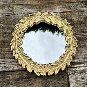May include: A round mirror with a decorative frame of leaves and flowers, finished in gold. The mirror is reflecting a blurred image of trees and sky.