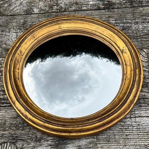 Puede incluir: Un espejo redondo con un marco dorado simple y adornado. El espejo refleja un cielo nublado.