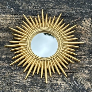 May include: A gold sunburst mirror with a circular reflective centre. The mirror features a textured starburst design with pointed rays. It is set against a weathered, dark wood surface. The mirror's diameter is approximately 20 cm.