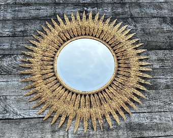 May include: A round, gold-colored sunburst mirror. The mirror has a circular reflective surface surrounded by ornate, radiating golden rays. The frame has a textured, detailed design, creating a striking decorative piece. The mirror is a decorative wall hanging.