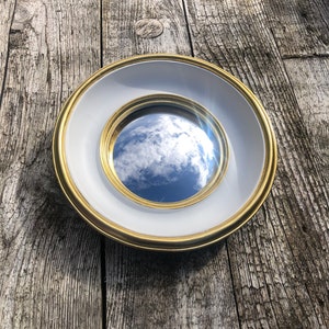 Può includere: Uno specchio rotondo con cornice bianca e bordo dorato. Lo specchio riflette un cielo blu con nuvole bianche.