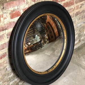 Op de afbeelding: Een ronde bolle spiegel met een zwart frame en een gouden binnenrand. De spiegel reflecteert een kamer met meubels en andere objecten.