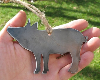 May include: A metal pig ornament with a rustic appearance. The pig-shaped decoration is made of gray metal and has a loop of twine for hanging. The ornament is held in a person's hand against a blurred green background.