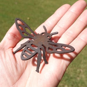May include: A flat, dark brown, rust-colored metal cutout in the shape of a bee. The insect figure features a stylized design with open, hollowed-out sections within the wings and abdomen, creating a silhouette that is held in the palm of a hand.